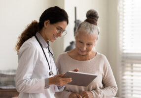 Happy female doctor and senior patient look at tablet screen discuss treatment or therapy on gadget. Smiling woman nurse and mature client use pad device discussing results in modern hospital.